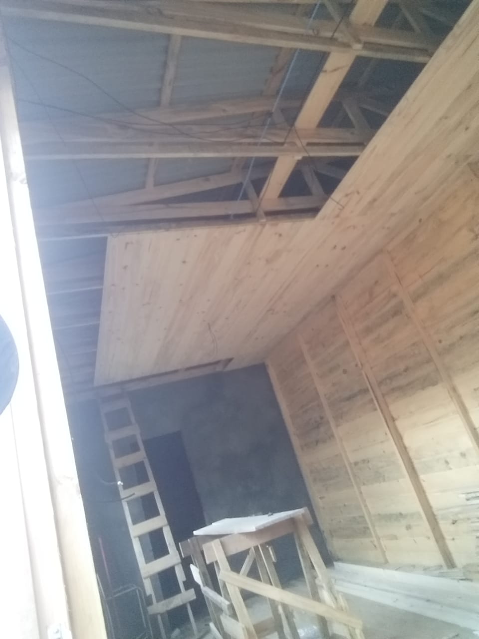 Interior de uma construção em andamento com estrutura de madeira, telhado de metal, escada de cimento e ferramentas de trabalho